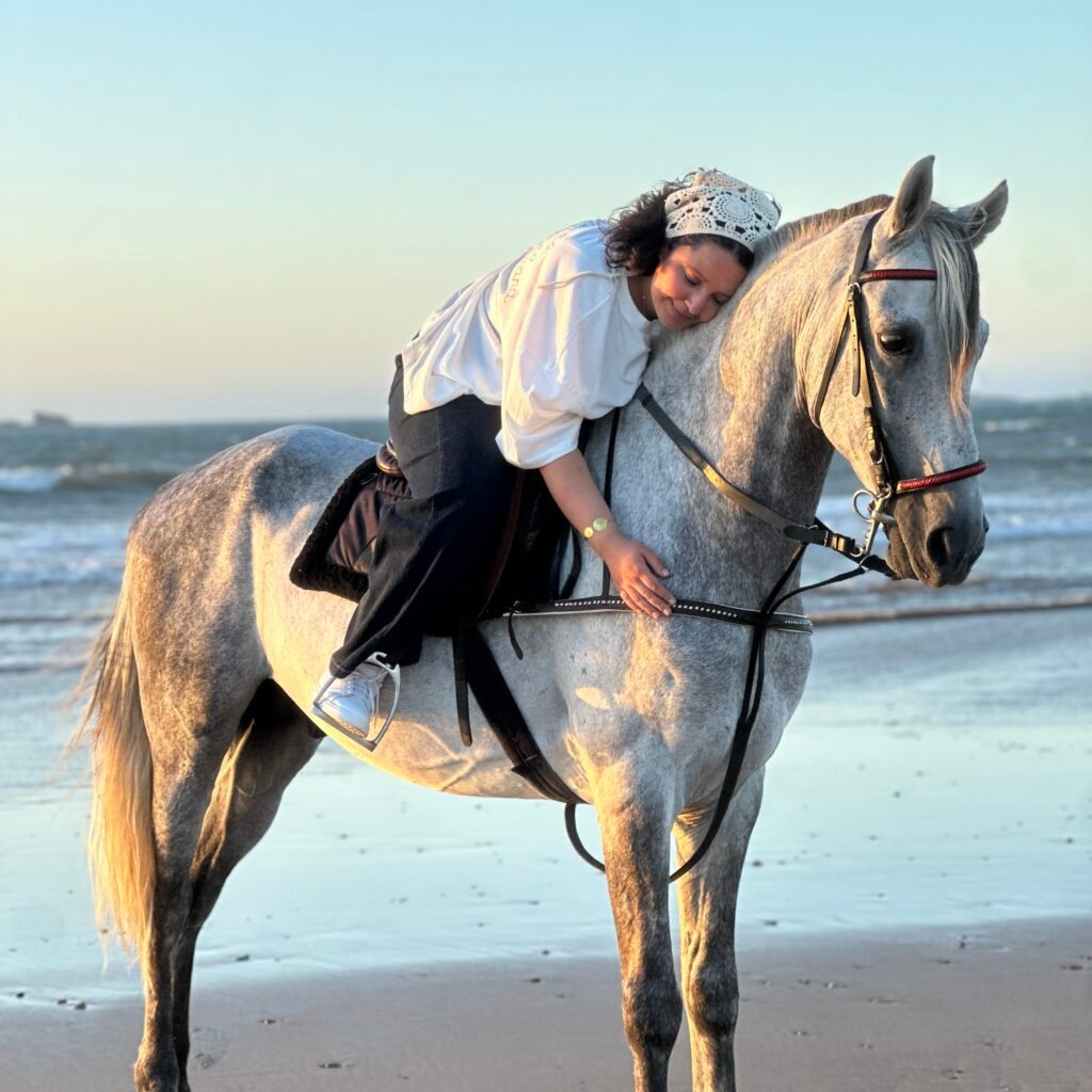 Female traveler leaning affectionately against her white horse on an beach at sunset
