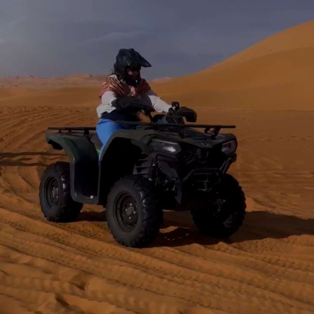 Person wearing a helmet riding an ATV quad bike across desert sand dunes