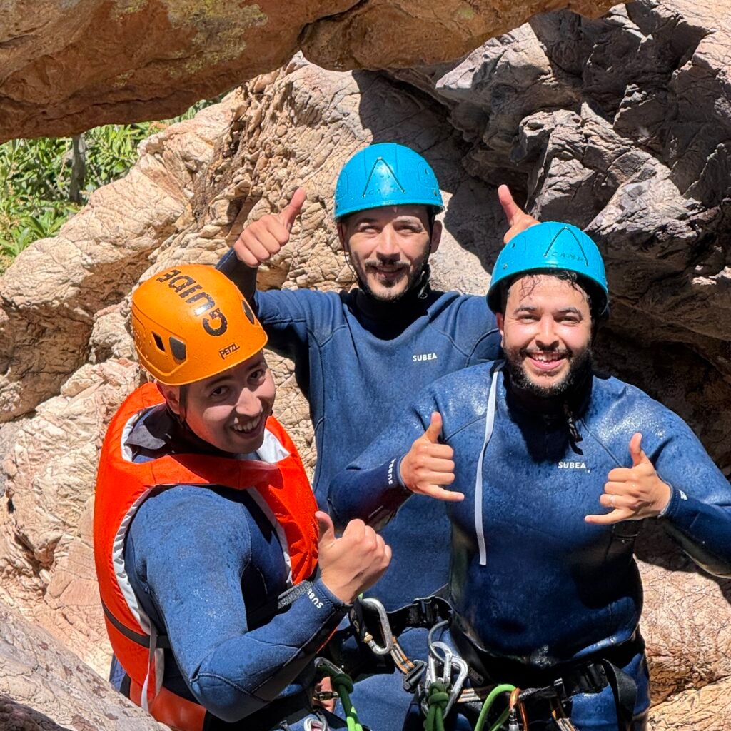 Three canyoneering guides wearing helmets and wetsuits gesturing thumbs-up in a rocky gorge in Morocco