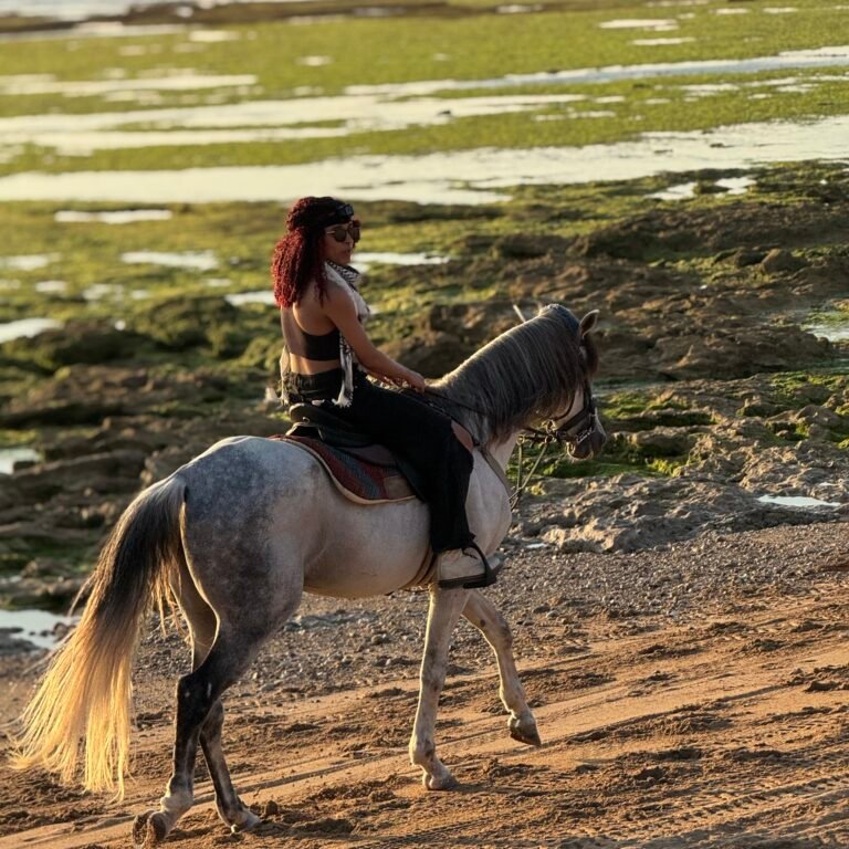 Woman with sunglasses riding a white horse near tide pools along the rocky coastline of Morocco