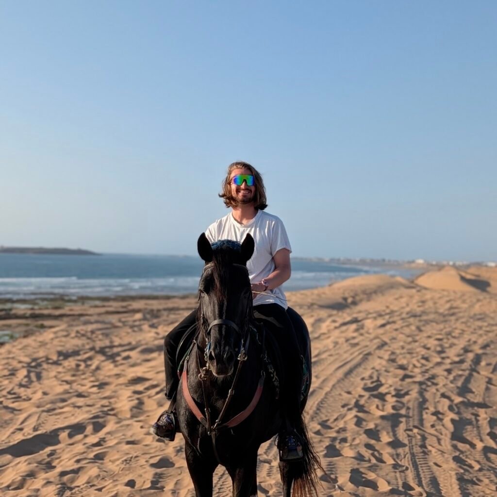Traveler with reflective sunglasses riding a black horse over sandy coastal dunes in Morocco