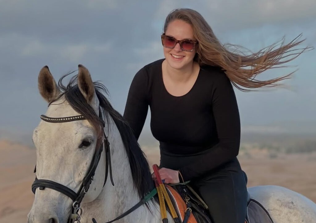 Female traveler in sunglasses riding a white horse across desert sand dunes in Morocco