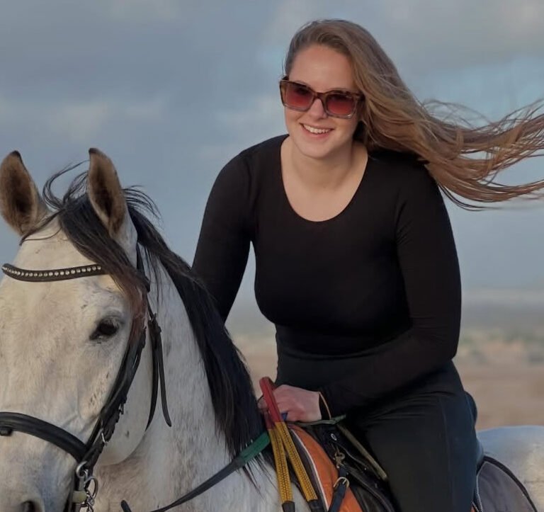 Female traveler in sunglasses riding a white horse across desert sand dunes in Morocco