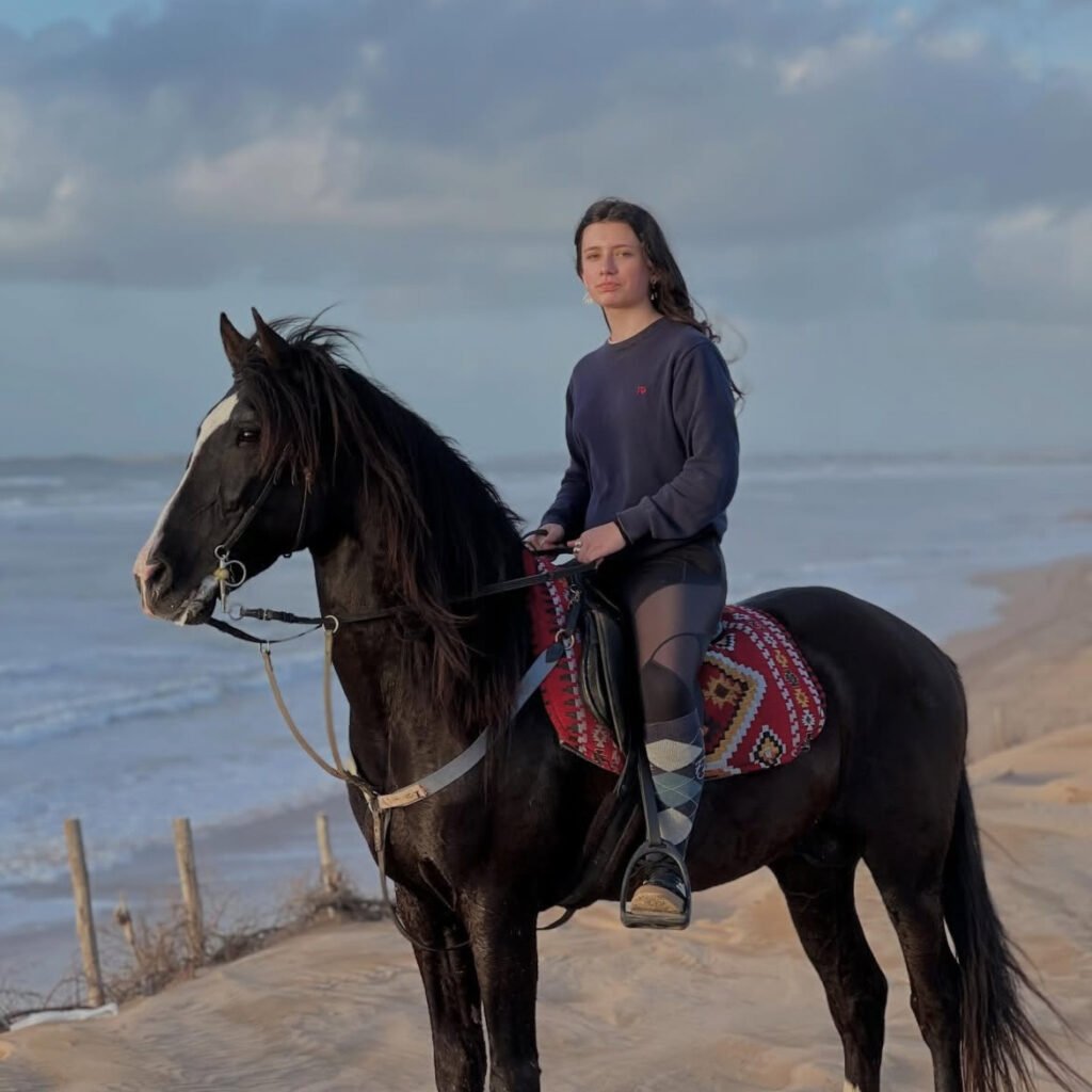 Female traveler riding a dark horse on a sandy dune overlooking the ocean coast in Morocco