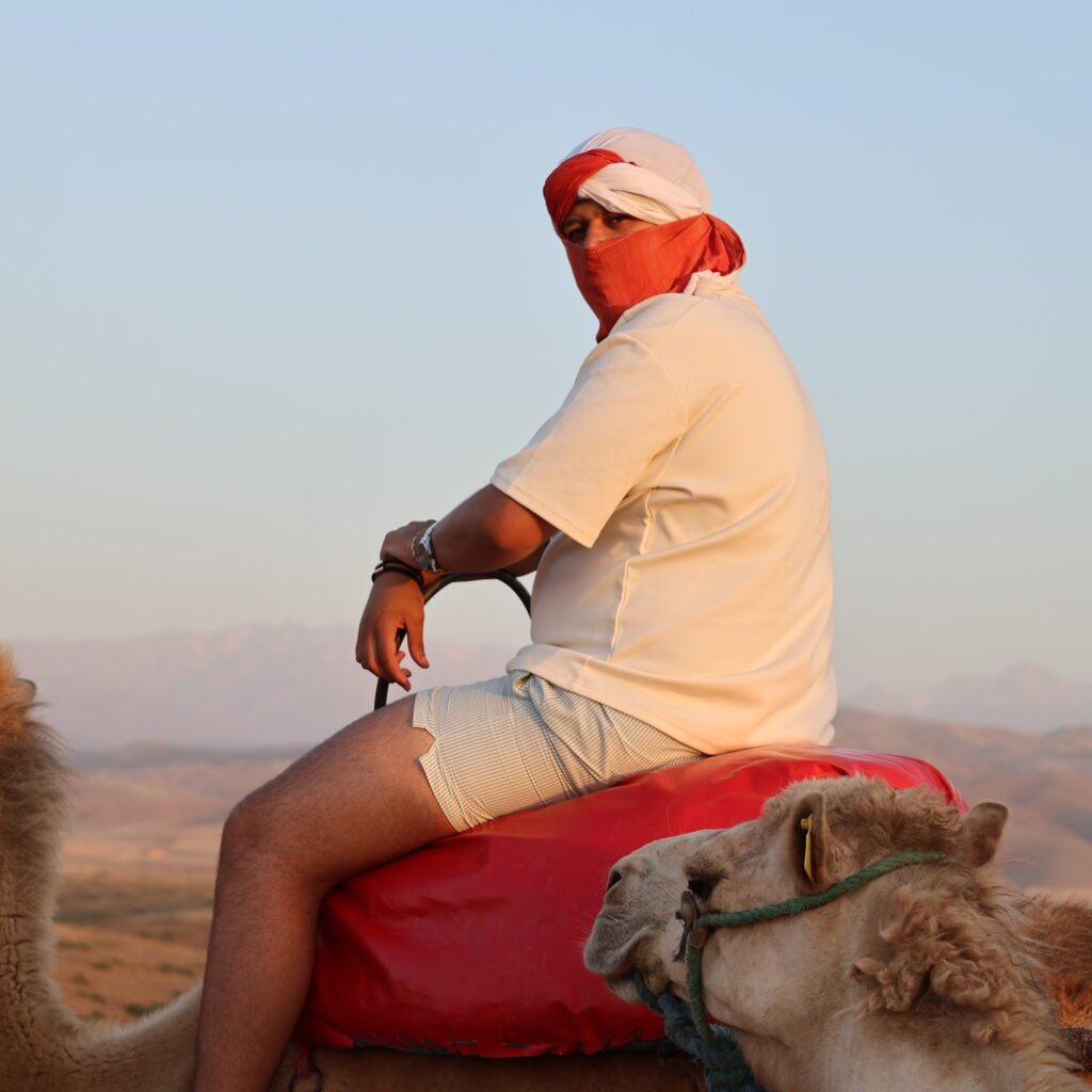 Male traveler wearing a red face scarf riding a camel during golden hour in Morocco