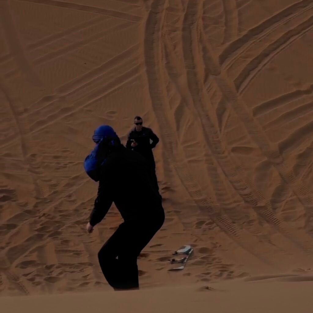 Person in a blue headscarf running up a sand dune during a desert sandboarding adventure tour