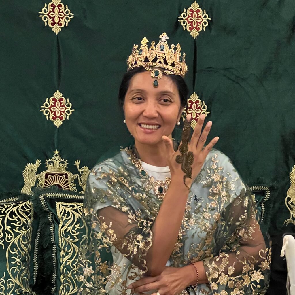 A woman wearing a crown and traditional embroidered garment smiling and showing a henna tattoo on her hand