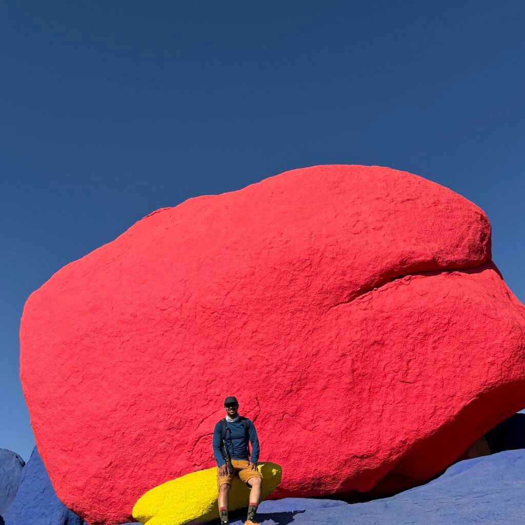 Traveler sitting on a painted yellow boulder in front of a giant red painted rock in Tafraoute, Morocco