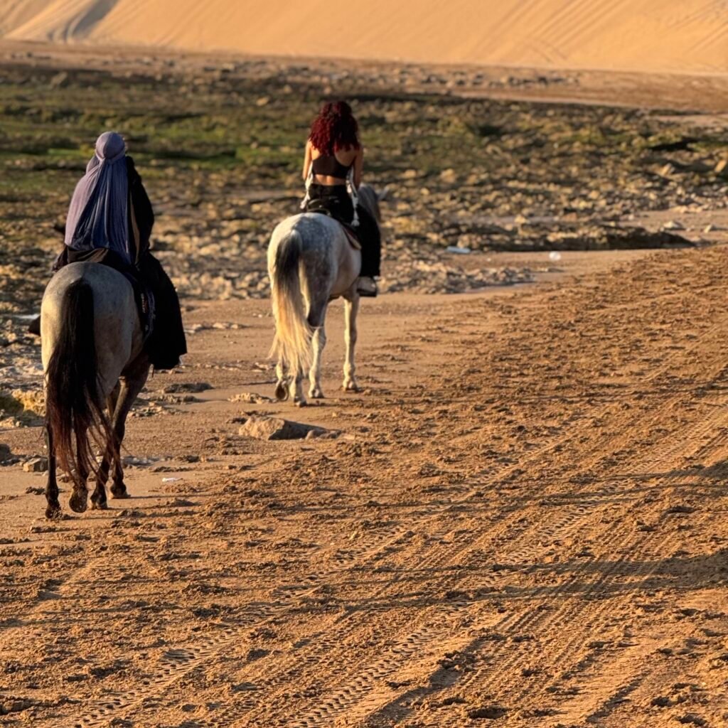 Two travelers horseback riding on a sandy beach path with desert sand dunes in the background in Morocco