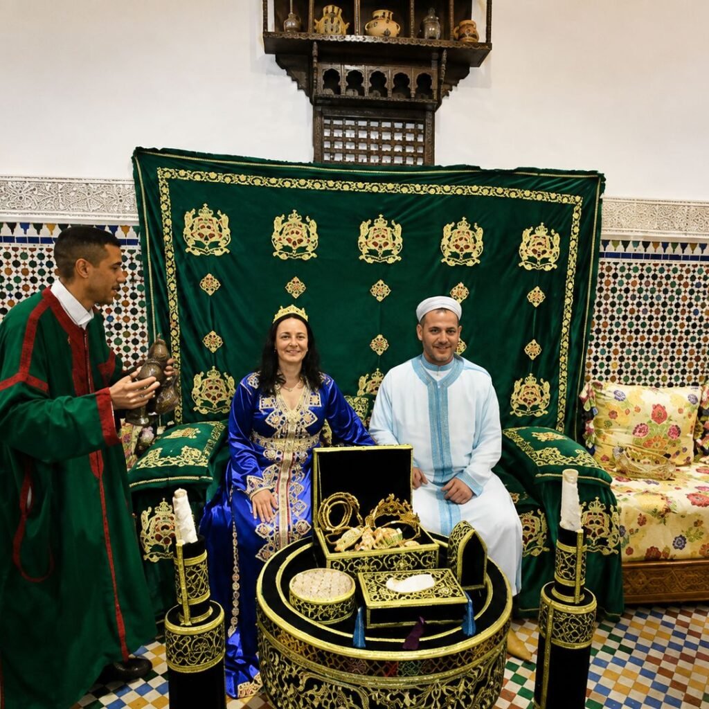 A couple posing on a decorative green throne chair during a traditional Moroccan wedding ceremony setup while a host stands nearby