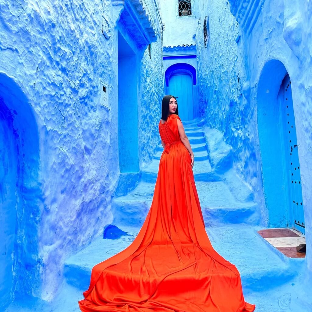 Woman in a flowing orange long-train dress posing on blue alley steps in Chefchaouen, Morocco