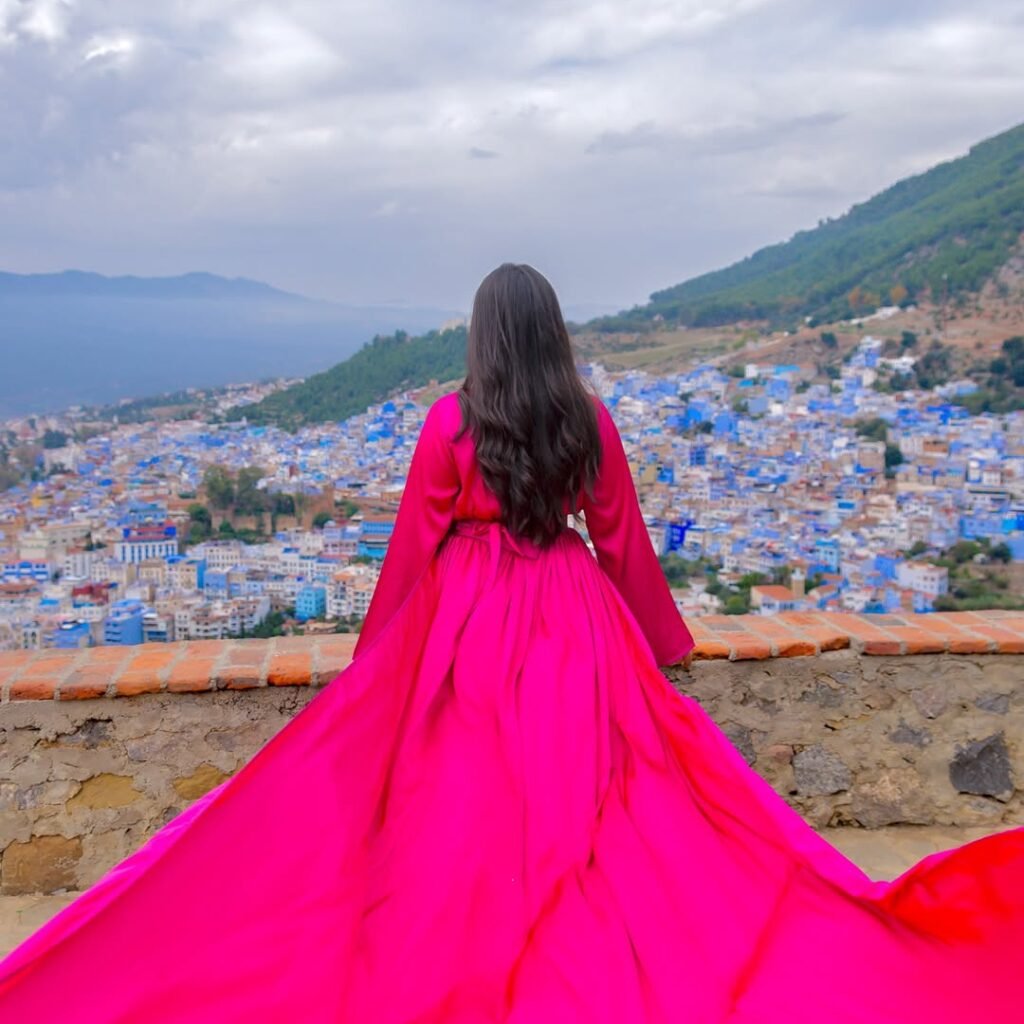 Rear view of a woman in a long magenta dress overlooking the hillside blue medina of Chefchaouen