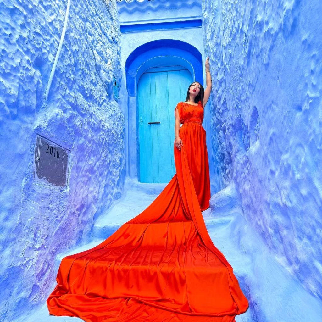 Female traveler posing in a long orange dress in a narrow blue street of Chefchaouen, Morocco