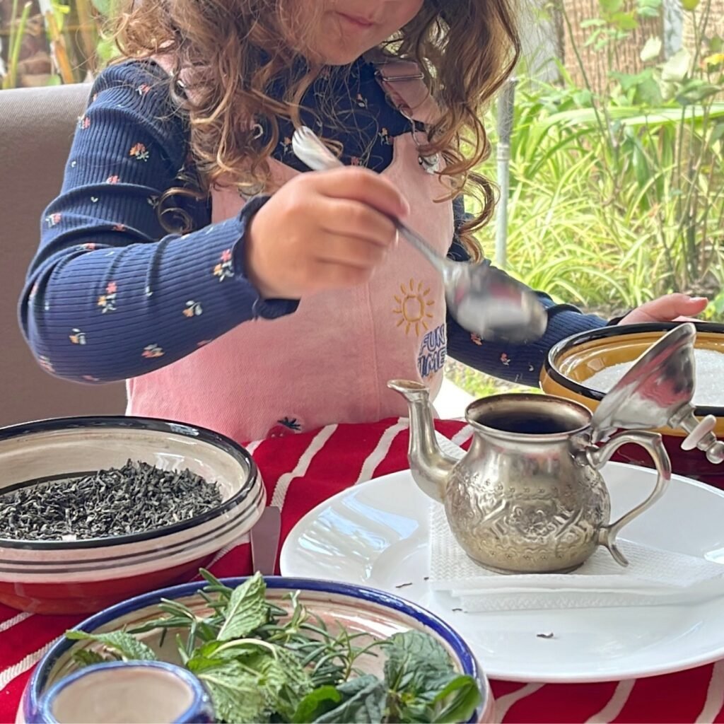 Young traveler making traditional Moroccan mint tea with fresh herbs and a silver teapot during a family workshop