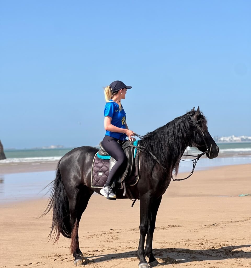 Female traveler in a blue shirt riding a black horse along a sandy beach in Agadir