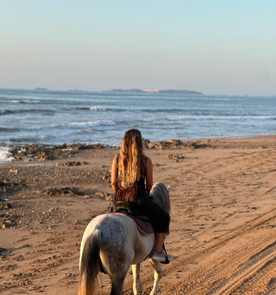 Rear view of a female traveler horseback riding a white horse along a beach path in Morocco