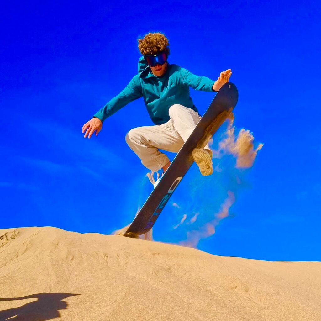 Man performing a sandboard jump trick off a desert dune peak against a blue sky