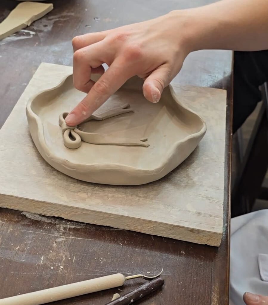 Person shaping a decorative clay ribbon onto a handmade dish in a pottery studio