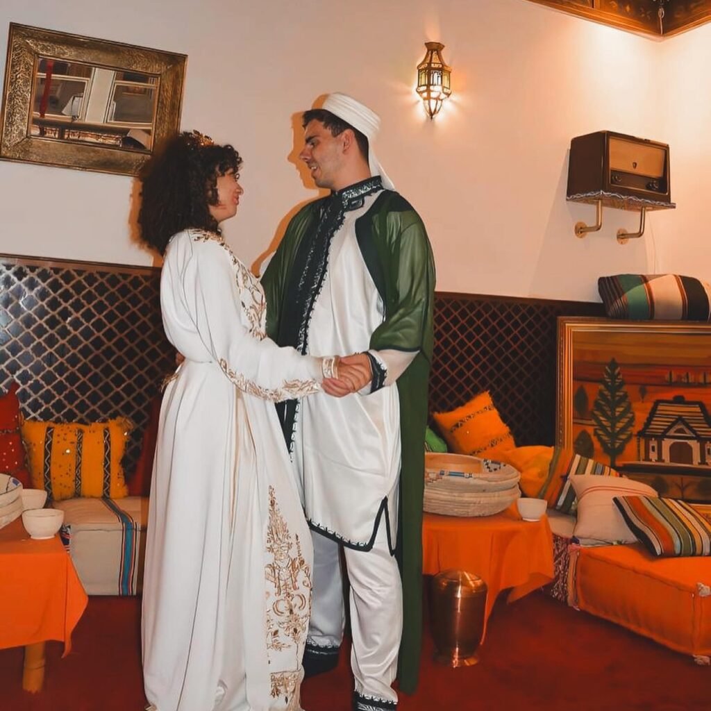 A couple holding hands in traditional attire inside a warmly lit room decorated with colorful cushions and textiles