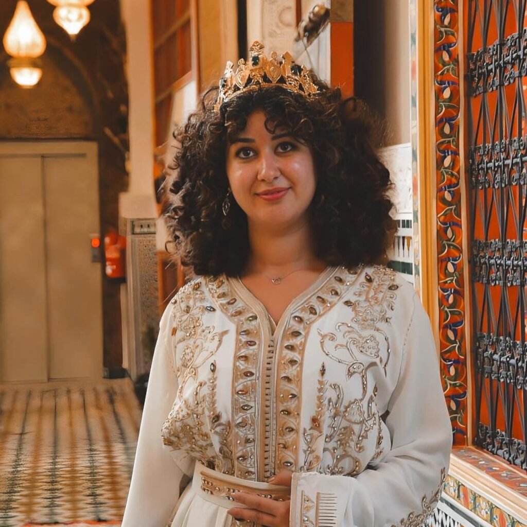 A traveler wearing an ornate crown and a white embroidered kaftan posing in a traditional venue