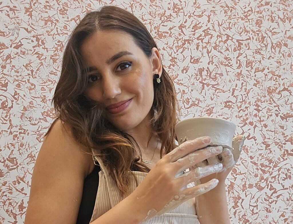 Smiling woman holding her freshly made ceramic cup covered in clay residue at a pottery studio