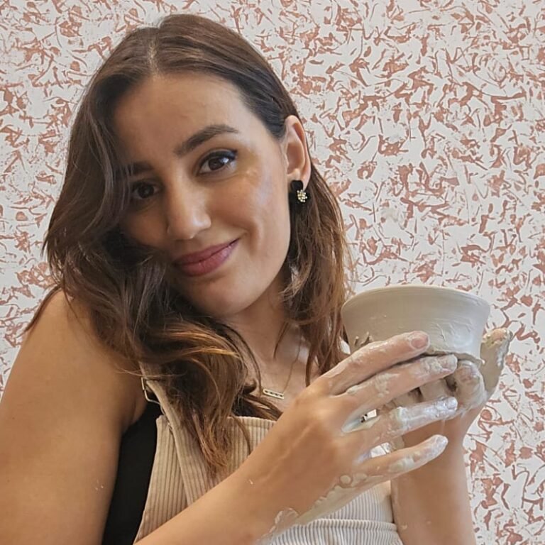 Smiling woman holding her freshly made ceramic cup covered in clay residue at a pottery studio